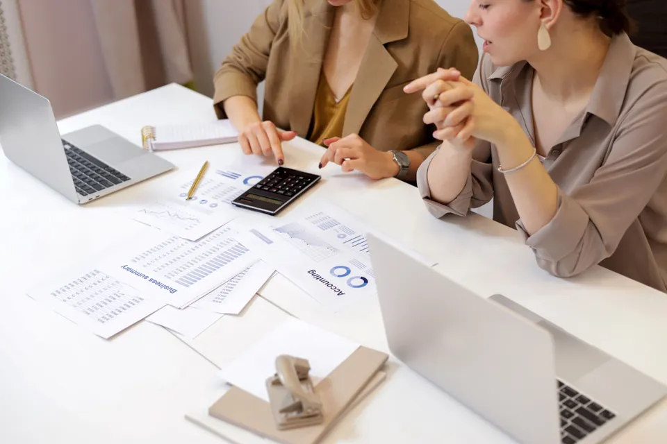 Two colleagues going through printed figures with a calculator, laptops open on the desk.