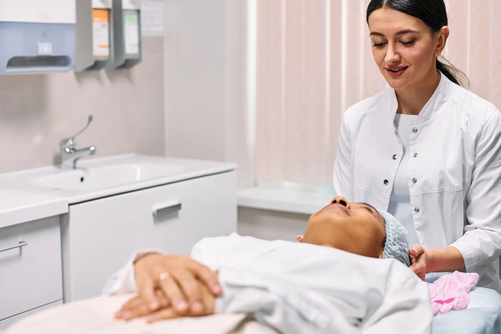 A practitioner giving a woman a skin treatment as she lies on a couch in a clinic room.