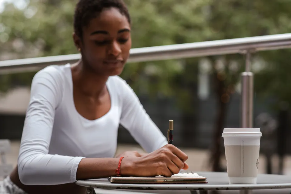 A woman writing in a notebook at an outdoor table, a takeaway coffee beside her.