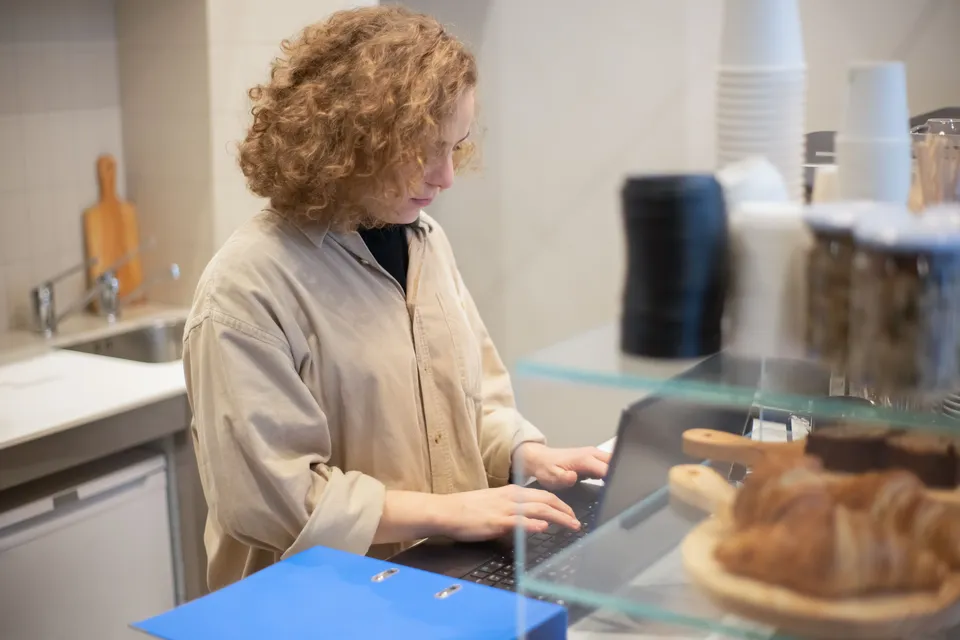 A café owner typing on a laptop behind the counter, pastries in the foreground.