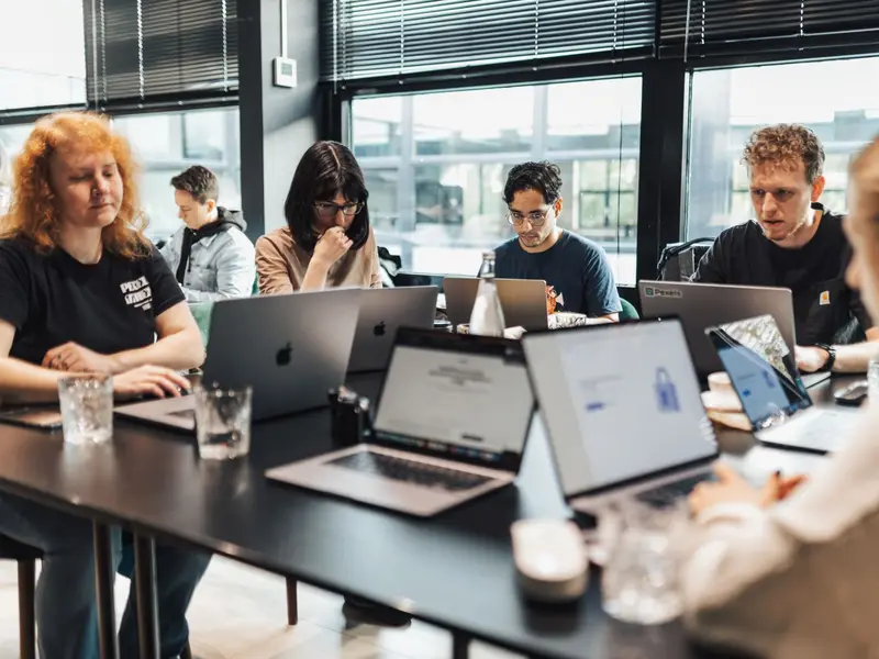 A group of people working on laptops around a long table in a bright office