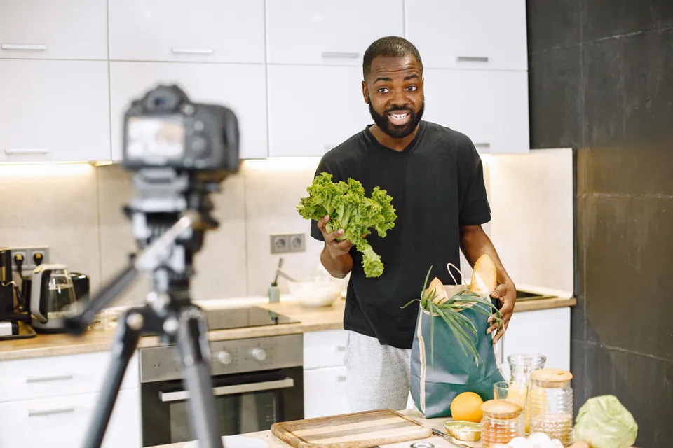 A man filming a cooking video in his kitchen, holding up a lettuce to the camera.
