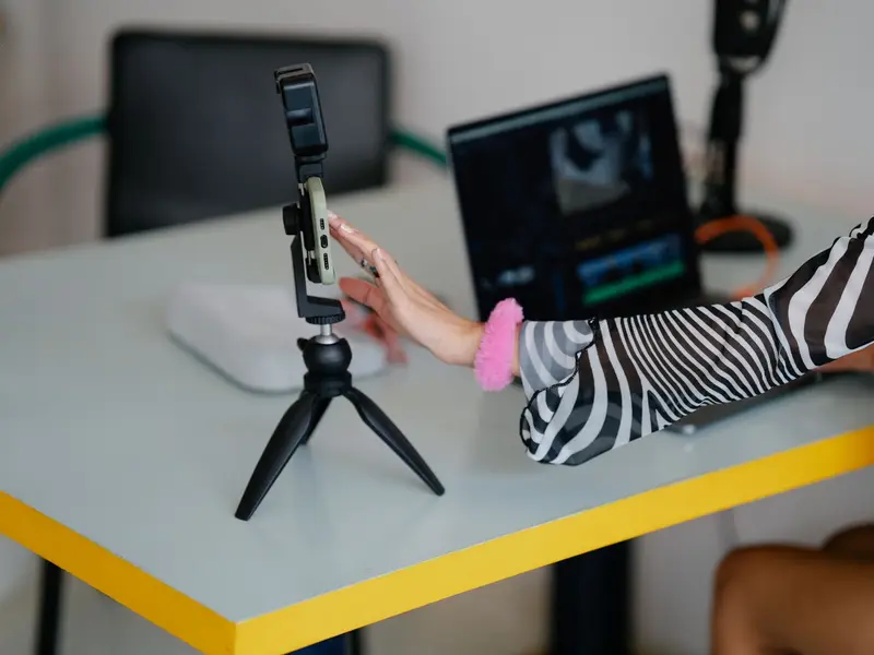 A person adjusting a phone on a small tripod beside a laptop showing a video edit
