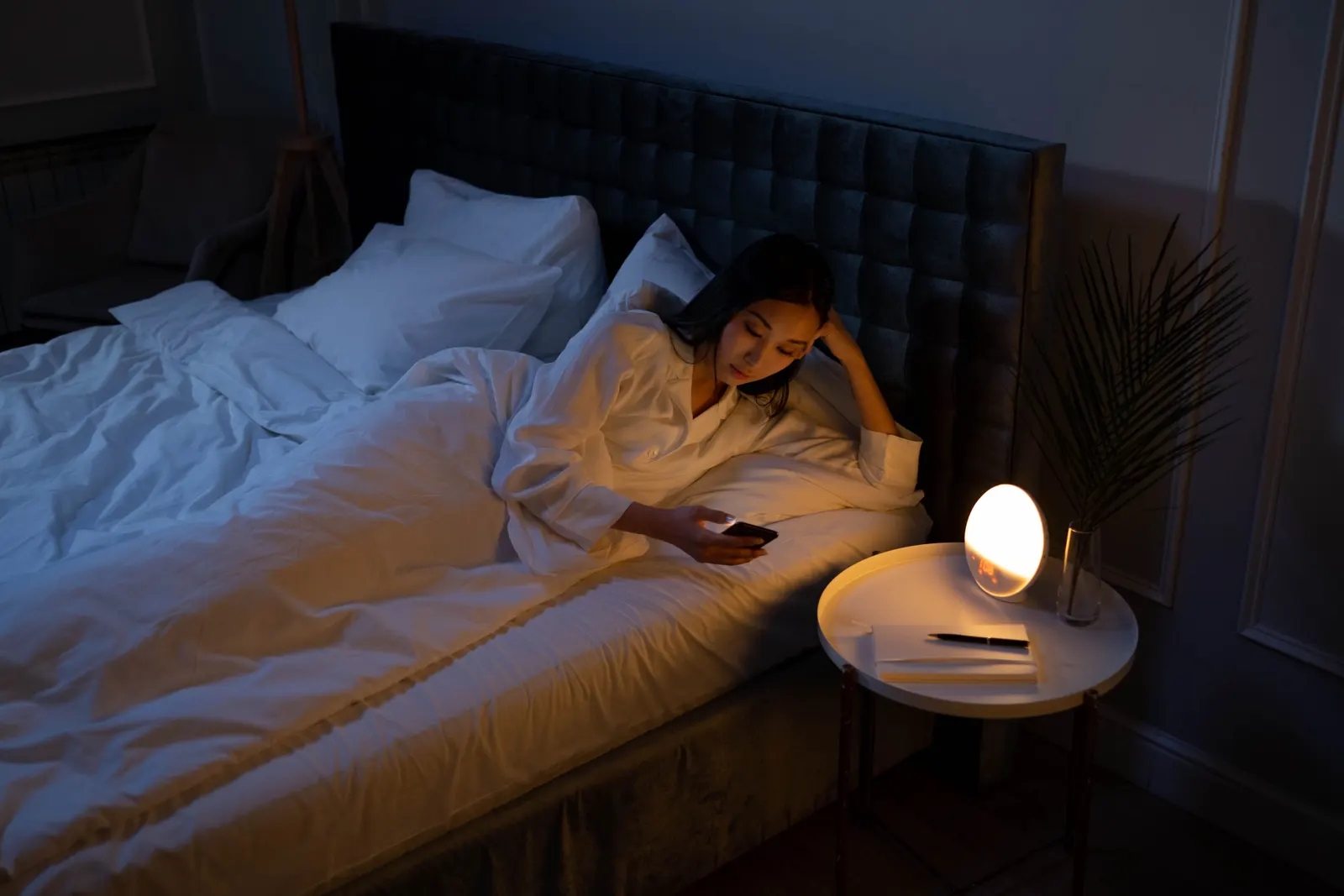 A woman lying in bed at night looking at her phone by the light of a bedside lamp.