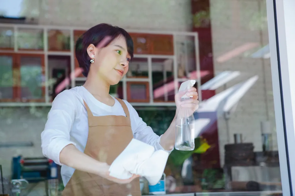 A shop worker in an apron cleaning the front window with a spray and a cloth.
