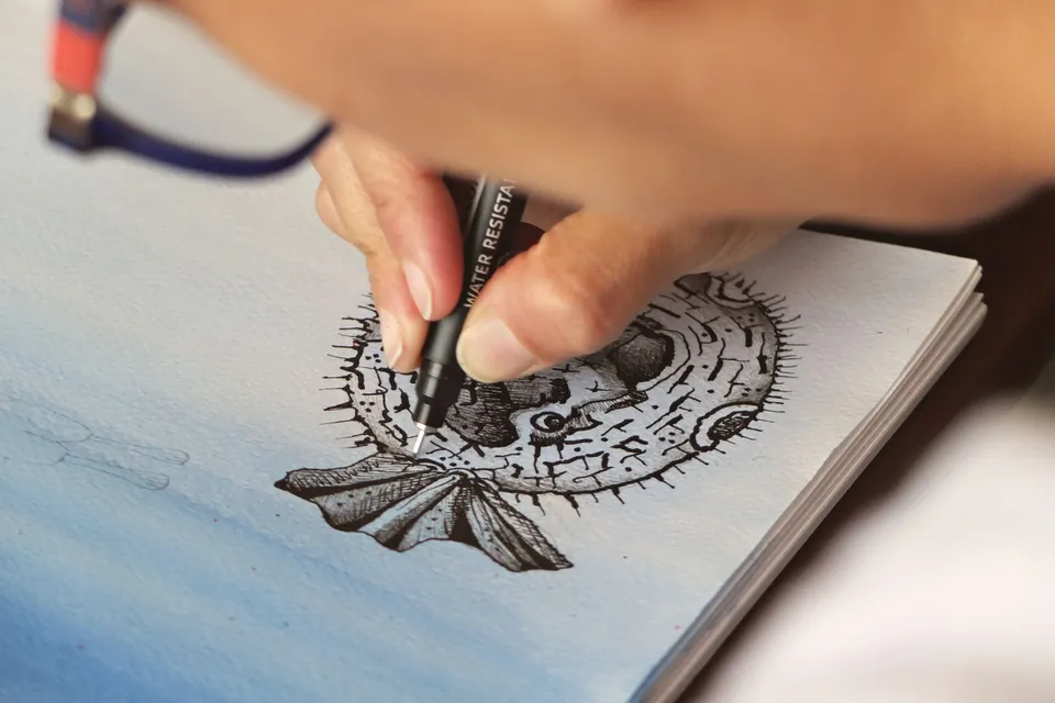 A hand inking a detailed drawing of a fish in a sketchbook.