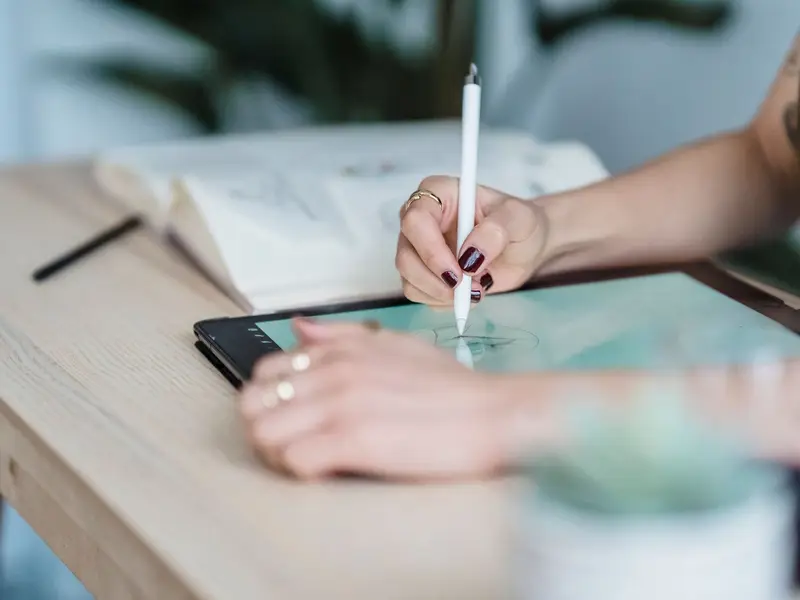 A hand drawing with a stylus on a tablet laid flat on a wooden table