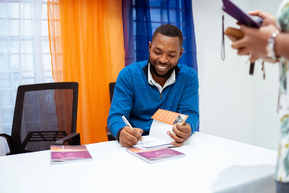 An author smiling as he signs a copy of his book at a table set out for a signing.
