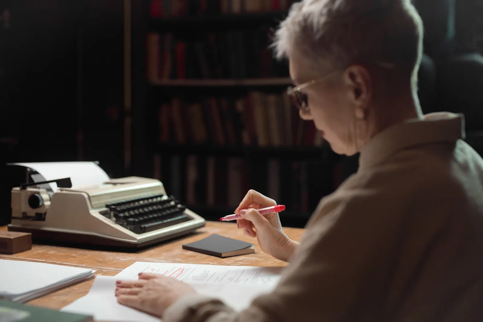 A writer reading over a manuscript at a wooden desk beside a typewriter.