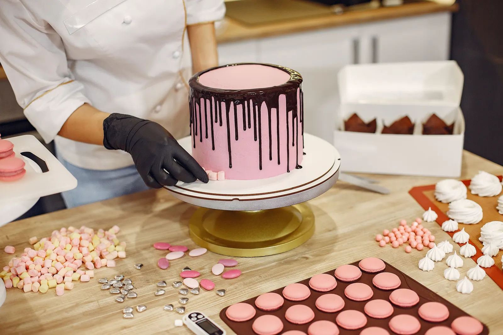 A gloved baker finishing a pink cake with a chocolate drip on a turntable, beside an open cake box and trays of macarons.