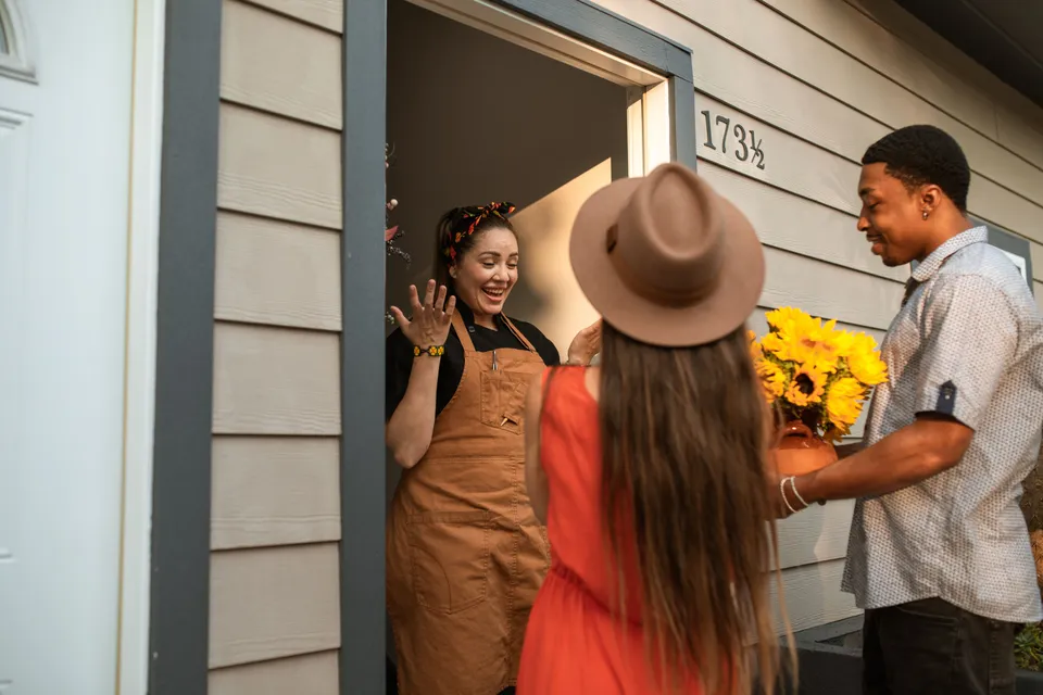 Guests arriving at a front door with a bunch of sunflowers, greeted by the host in an apron.