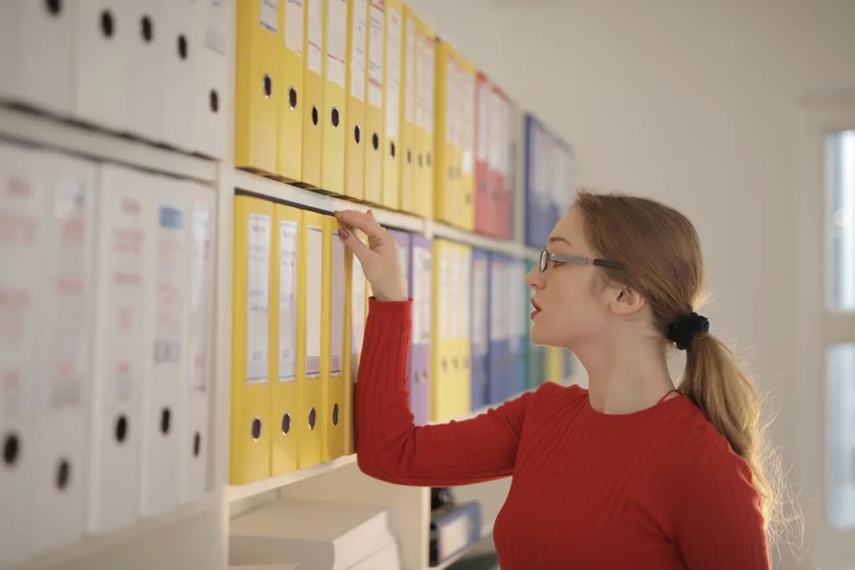 A woman reaching for a folder on a wall of colour-coded binders in an office.