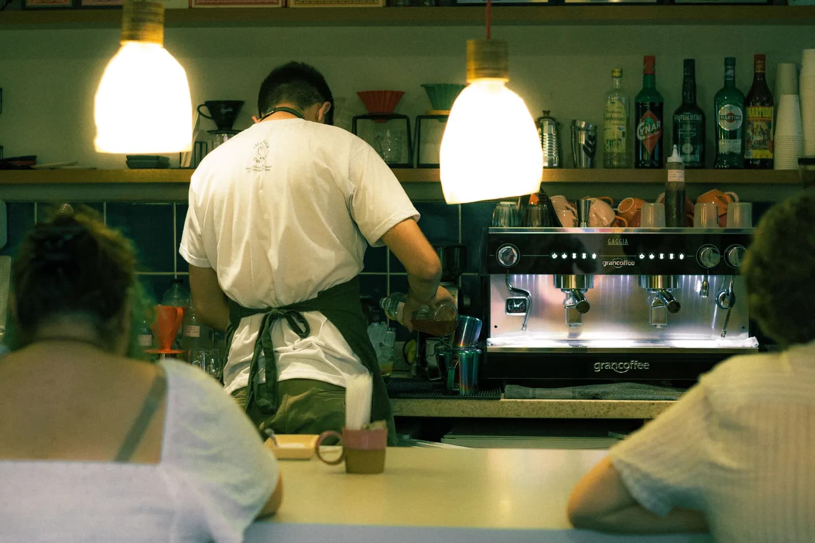 A barista working at the espresso machine behind a café counter, seen from behind two customers waiting on stools.