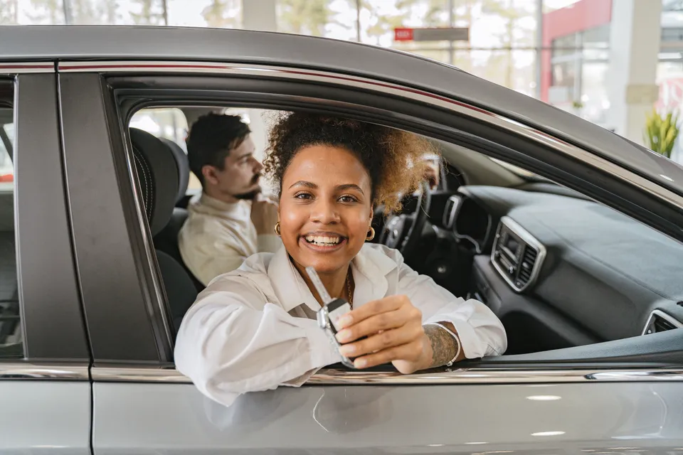 A smiling driver sitting in a new car and holding up the keys, a passenger beside her.