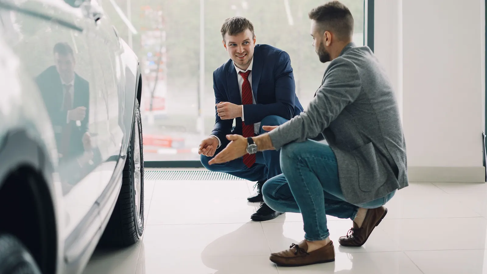 A salesman and a customer crouched beside a car in a showroom, talking it over.