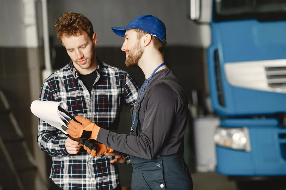 A mechanic going through a clipboard with a customer in the workshop, a lorry behind them.