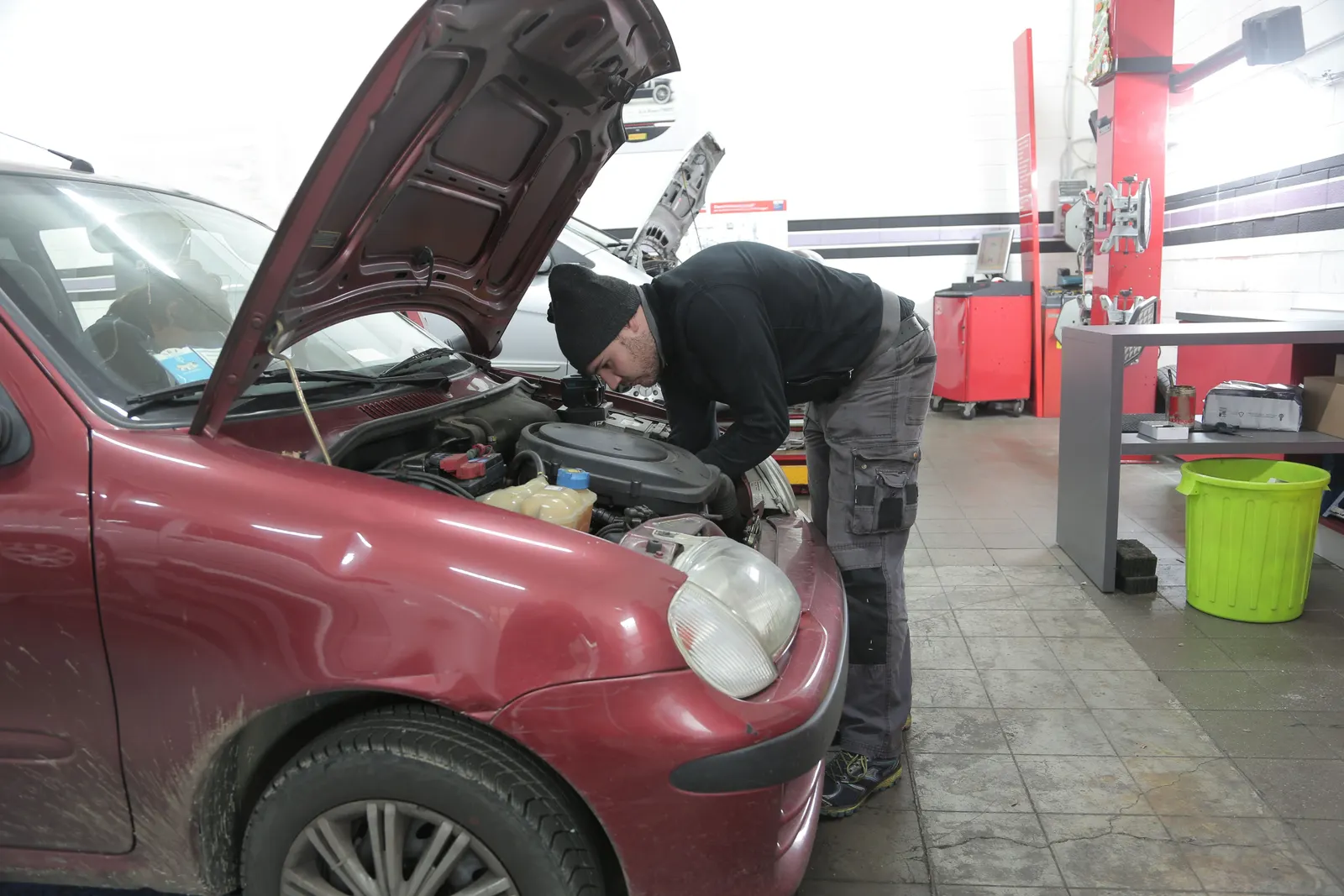 A mechanic leaning into the engine bay of a red car with its bonnet up in a workshop.