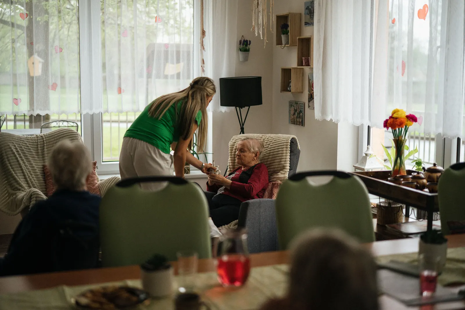 A carer kneeling beside two elderly residents taking tea in a care home lounge.