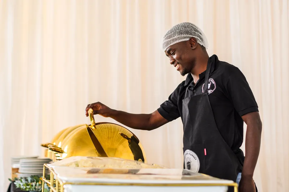 A caterer in a hairnet lifting the lid of a gold serving dish at a buffet.