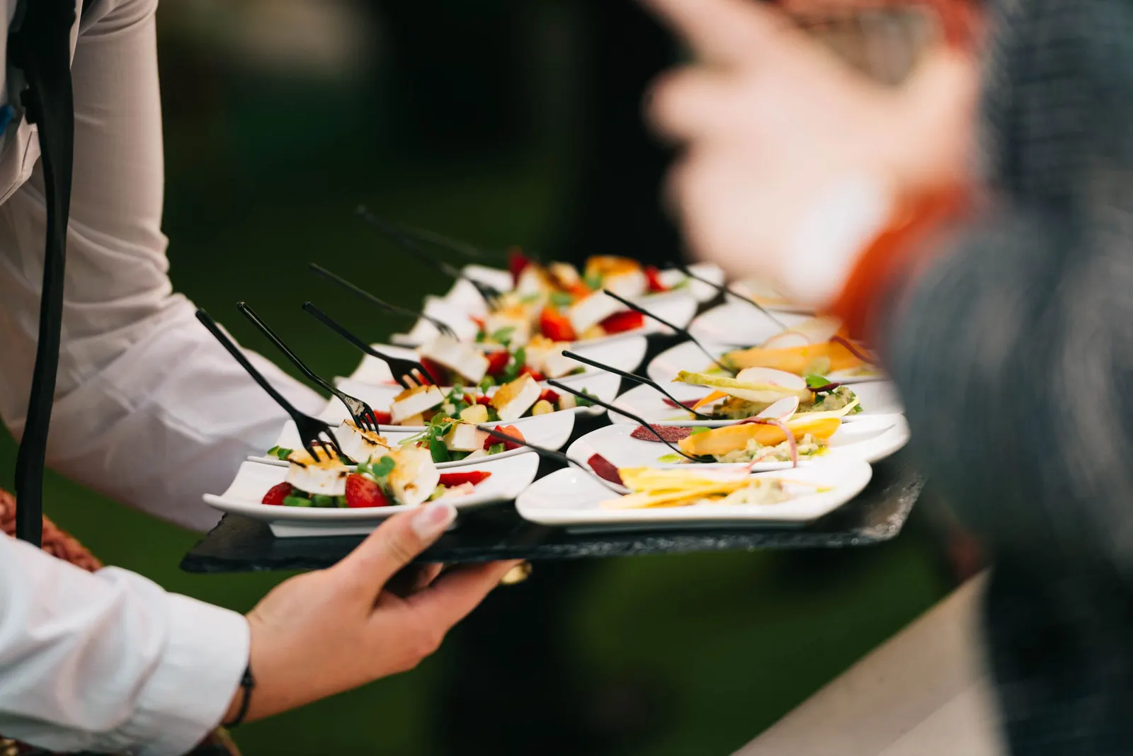 A server holding out a slate tray of small plated starters, each with its own fork, to guests at an outdoor event.