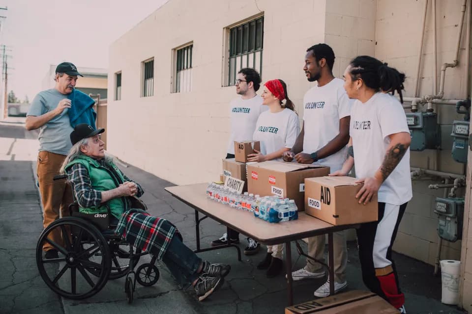 Volunteers handing out boxed supplies from a table outdoors, one person waiting in a wheelchair.