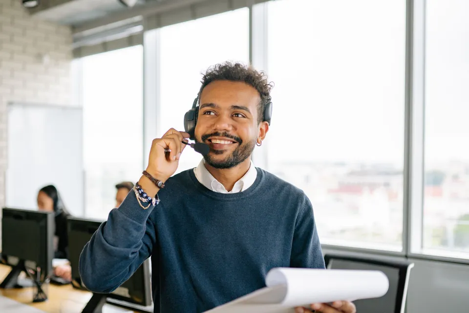 A support agent in a headset smiling as he takes a call, a clipboard in his hand.