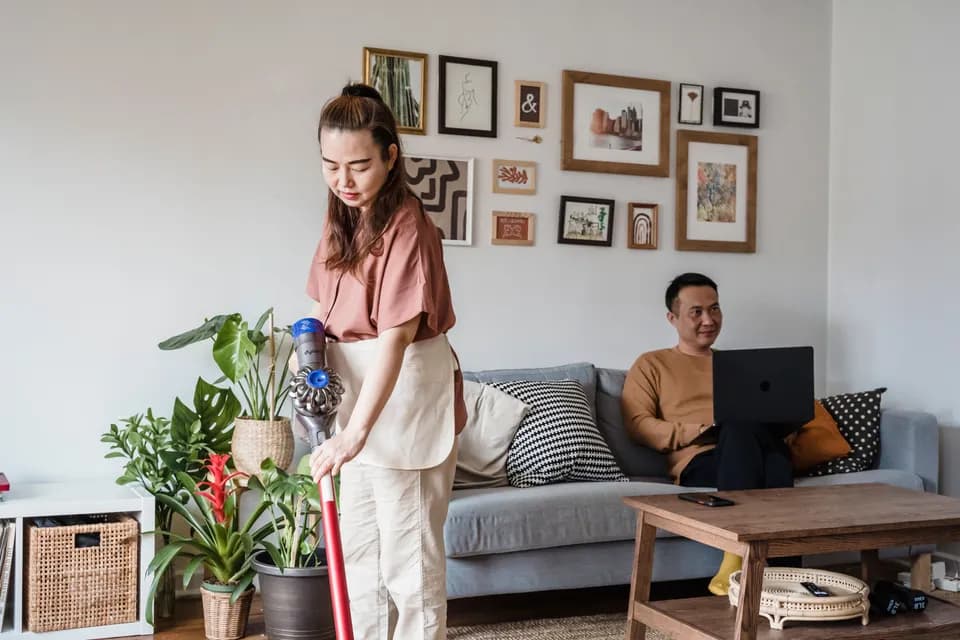 A cleaner working through a living room while the homeowner sits on the sofa with a laptop.