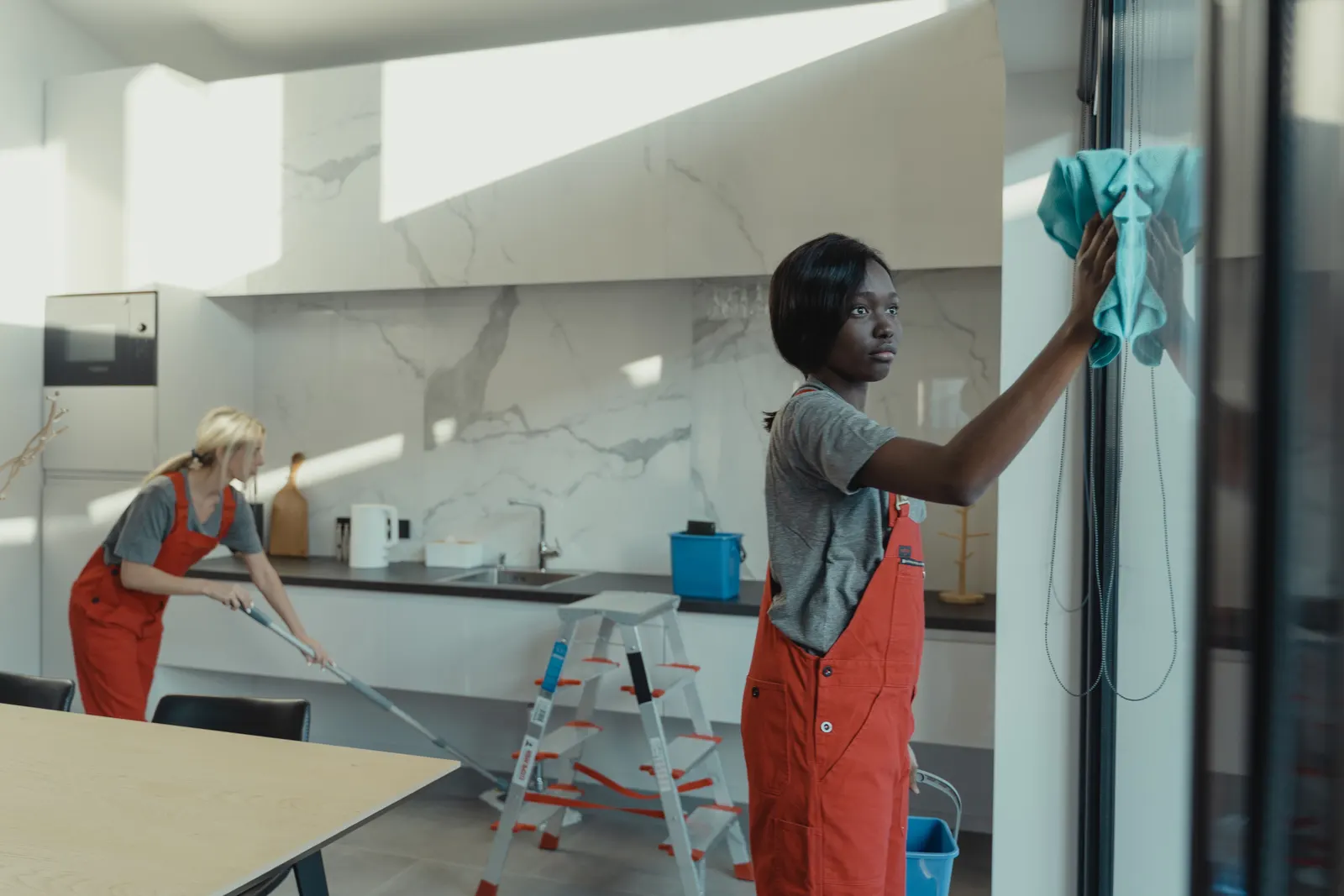 Two cleaners working through a modern kitchen, one wiping the glass and one mopping the floor.