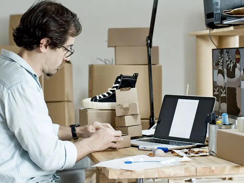 A man packing a customer order at his desk beside a laptop and stacked boxes