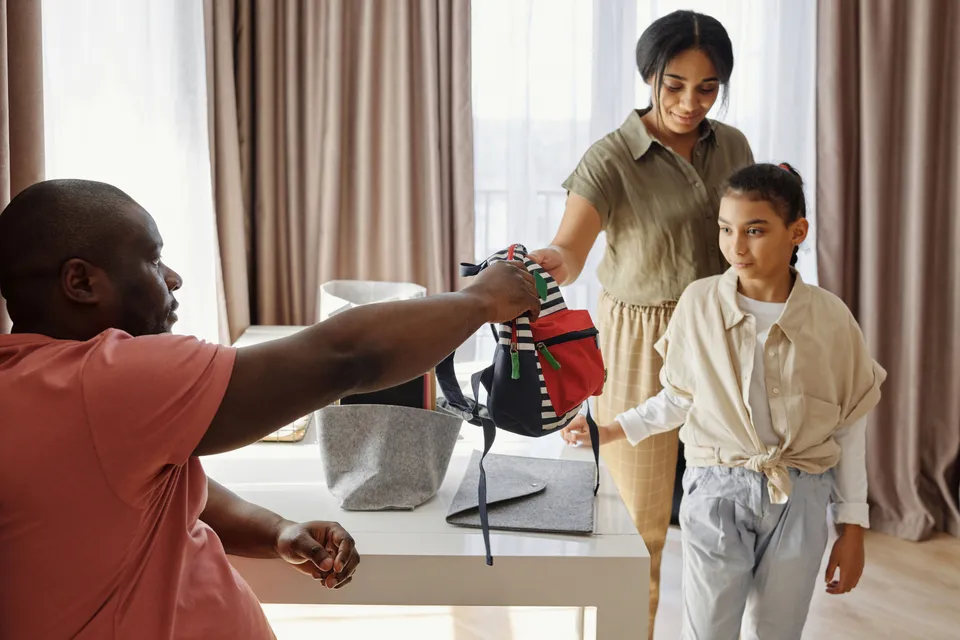 A parent handing over a child's backpack at the door as the child gets ready to leave for school.