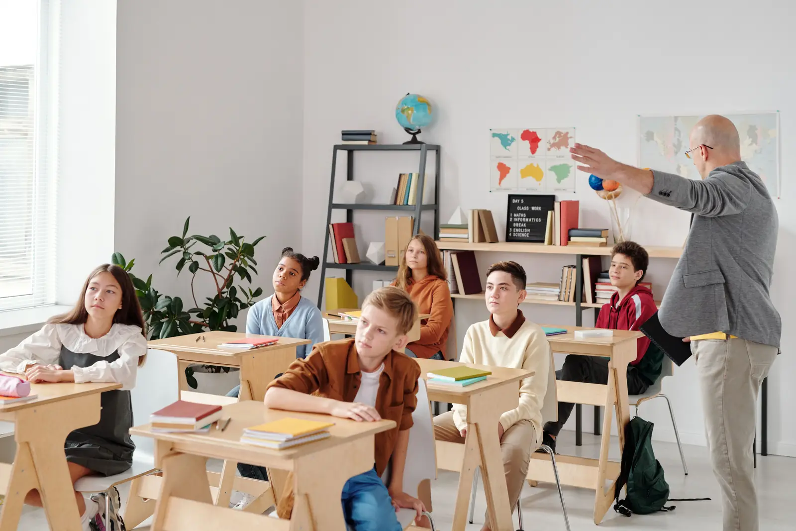 A teacher gesturing to the board while a class of teenagers listens from their desks.