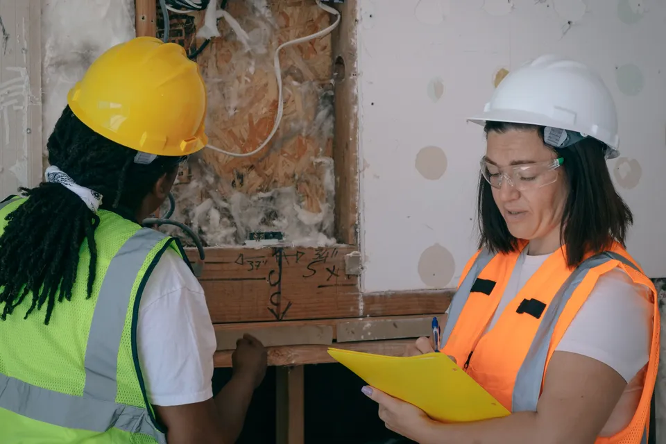 Two electricians in hard hats inspecting exposed wiring, one taking notes on a clipboard.