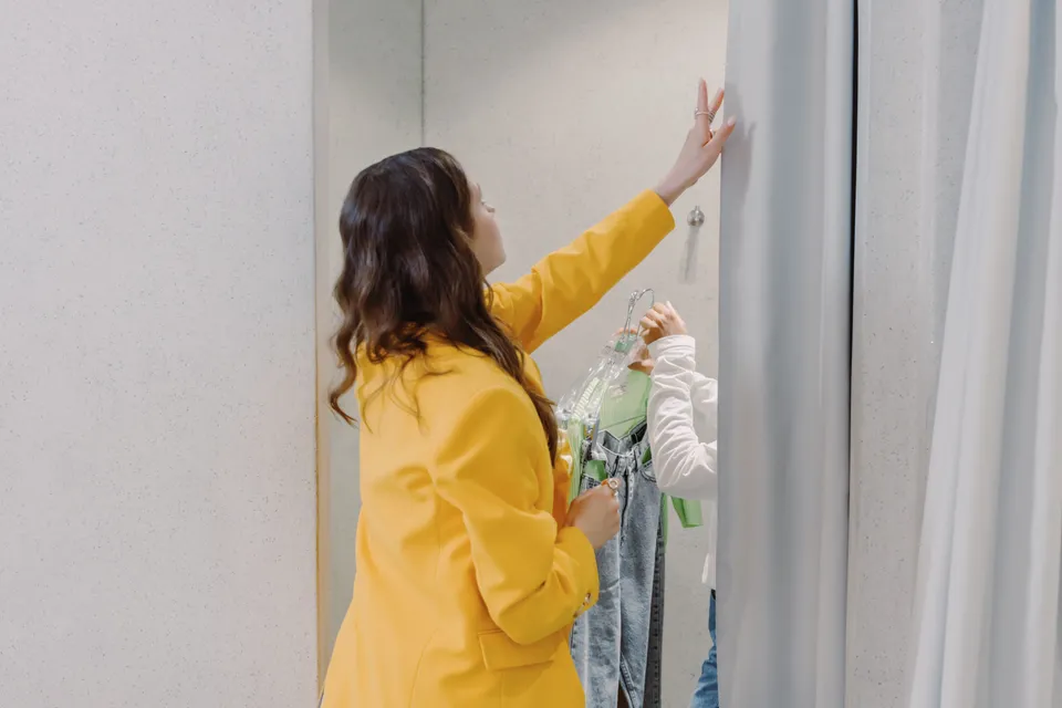 A woman in a fitting room reaching up to hang a garment while trying on clothes.