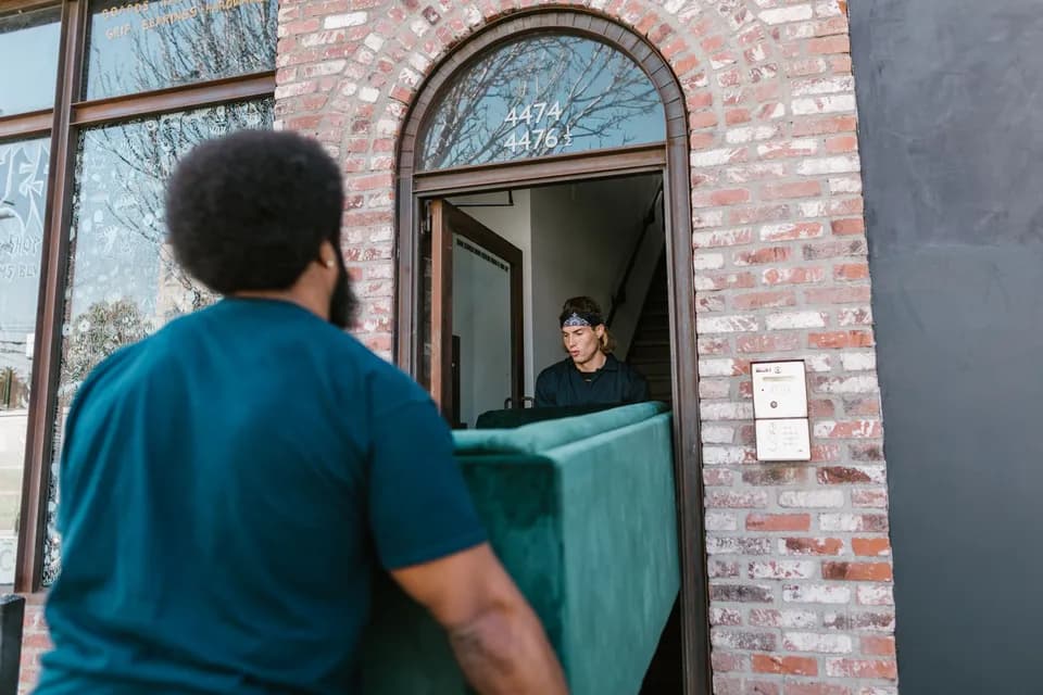 Two delivery men carrying a green sofa in through the front door of a brick house.