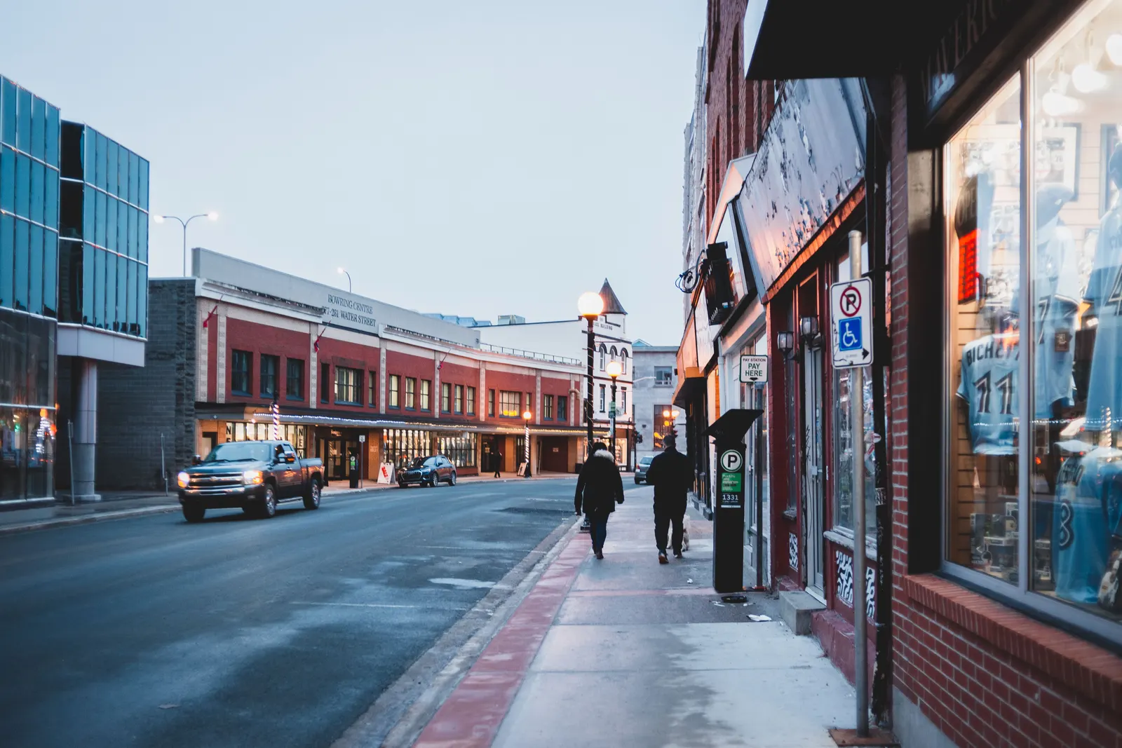 A high street of small shopfronts at dusk, with people walking past the windows.