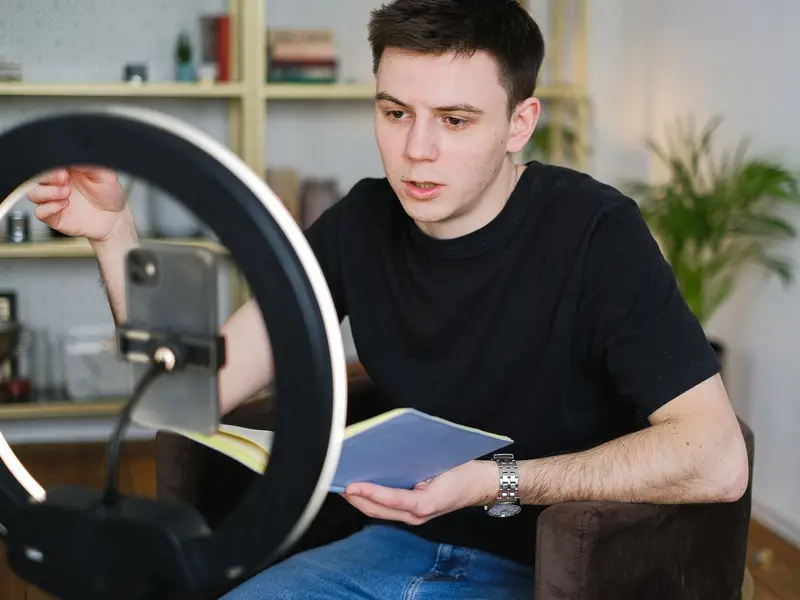 A young man recording himself on a phone mounted in a ring light, notebook in hand