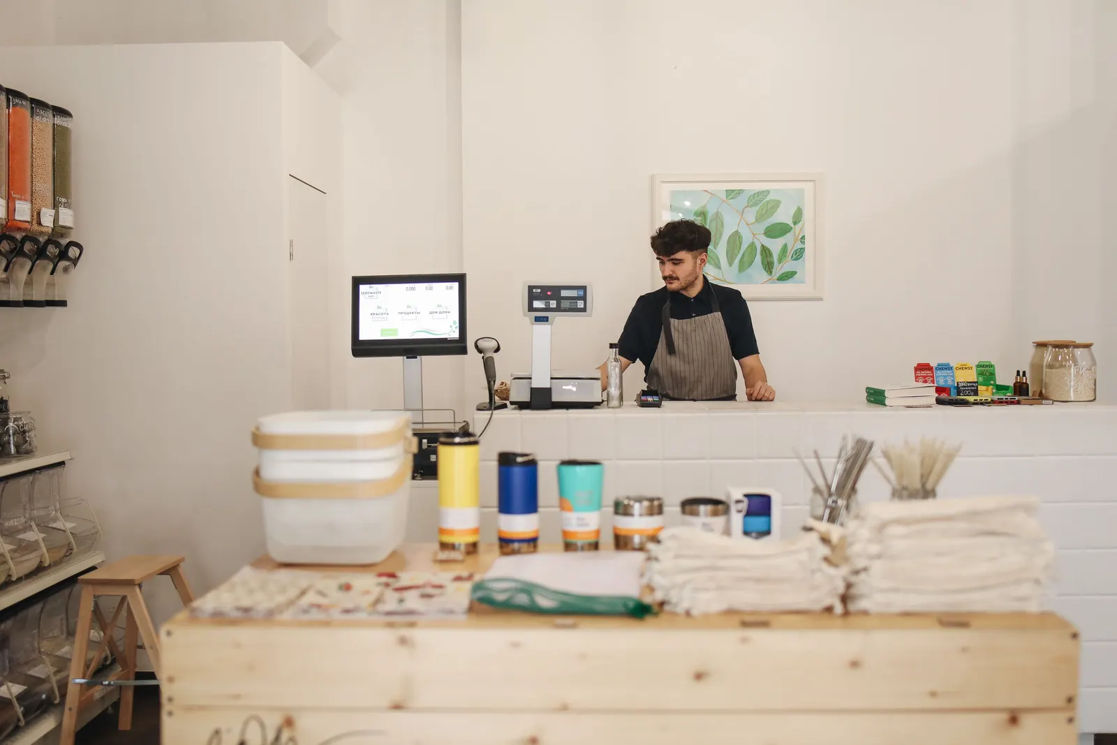 A young shop owner standing behind the counter of his own small shop, jars and goods on display.