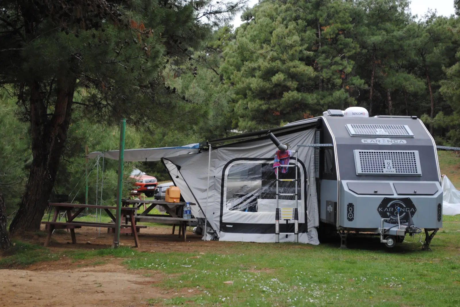 A caravan with its awning pitched among trees at a campsite, beside a wooden picnic table.