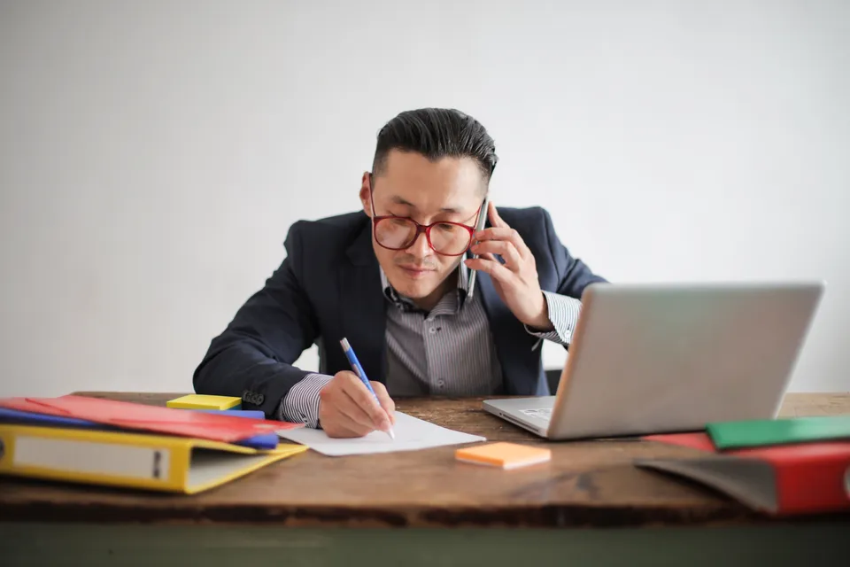A broker on the phone at his desk, taking notes beside an open laptop and folders.