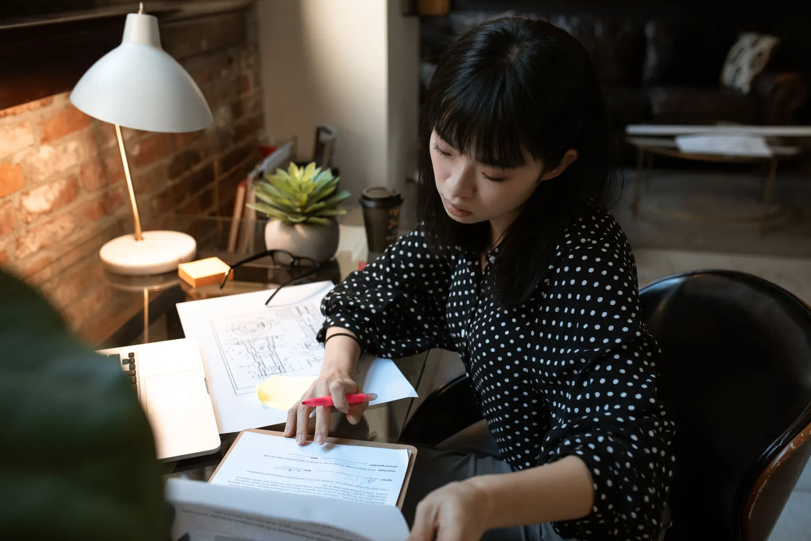 A woman working through papers and a notebook at a desk under a lamp, late in the day.
