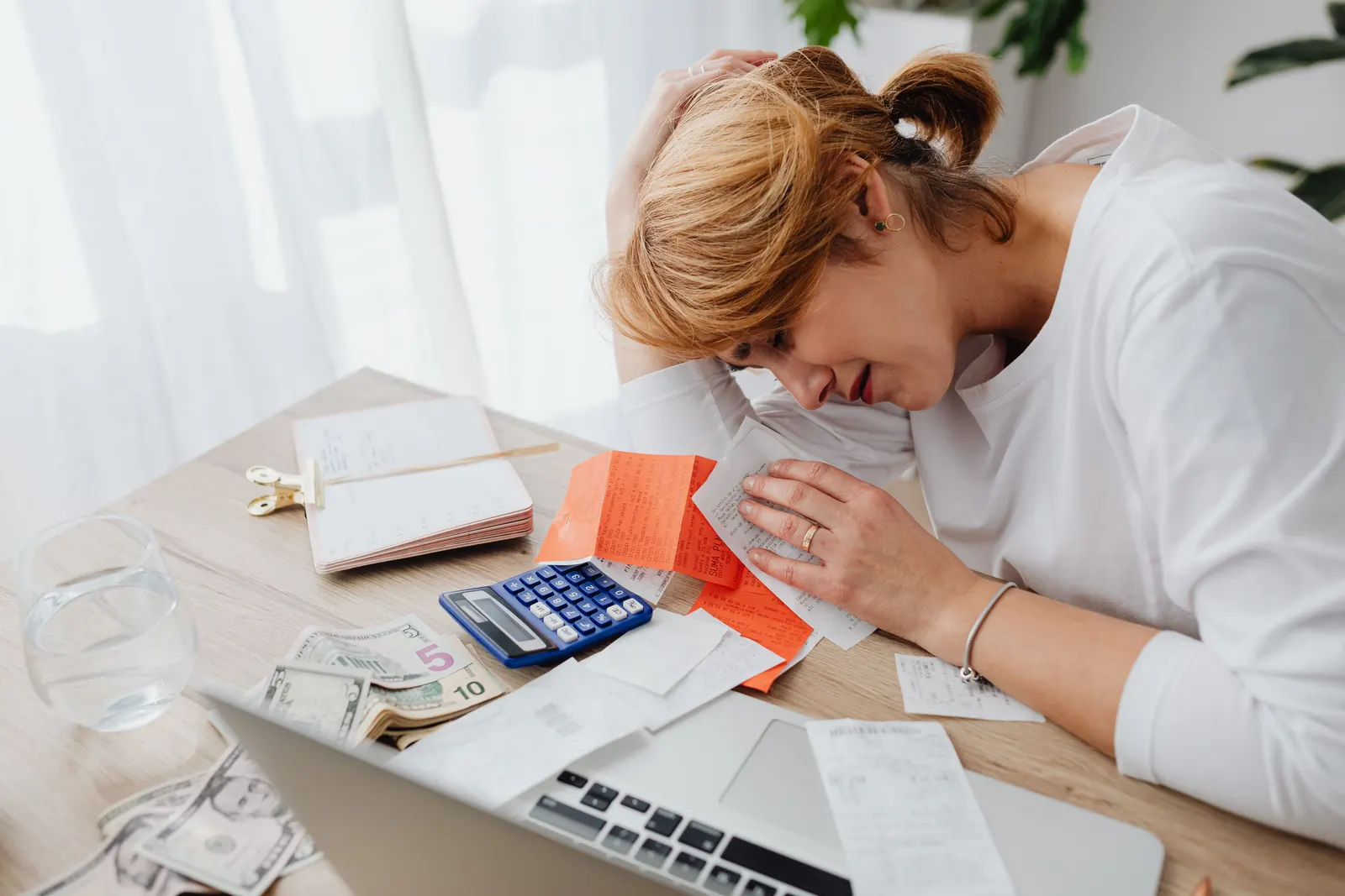 A woman resting her head in her hand over a spread of receipts, a calculator and cash.