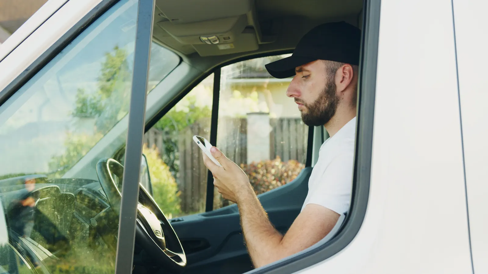 A man in a cap checking his phone in the driver's seat of a parked van.