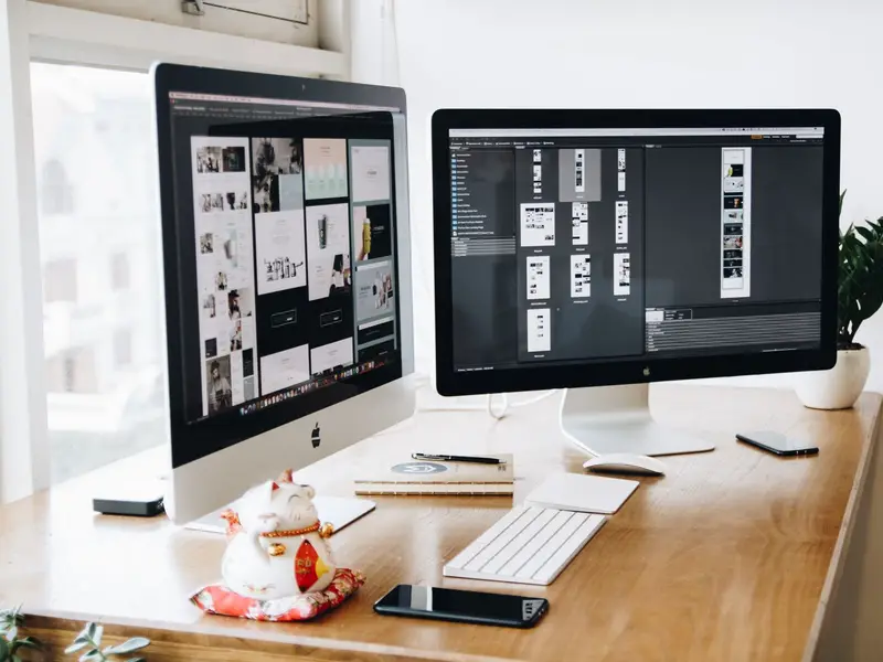 Two desktop monitors on a wooden desk showing web page layouts side by side