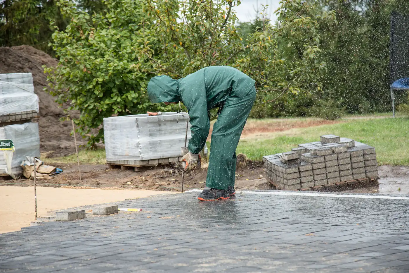 A worker in waterproofs laying paving stones in a garden on a wet day.