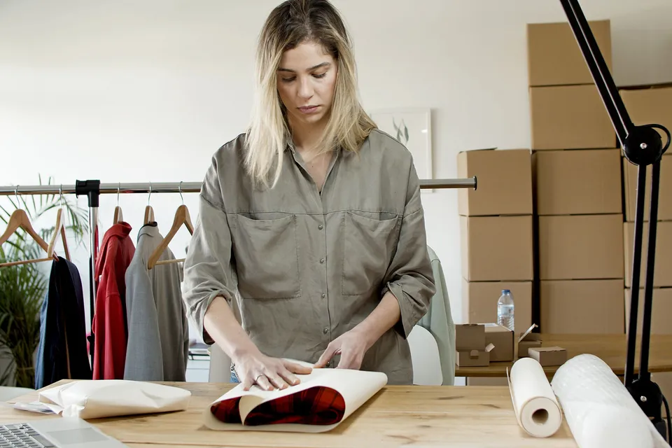A woman wrapping clothes in paper at a packing table, boxes stacked behind her.