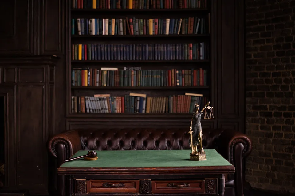 A dark study with shelves of books, a gavel and a figure of Justice on the desk.