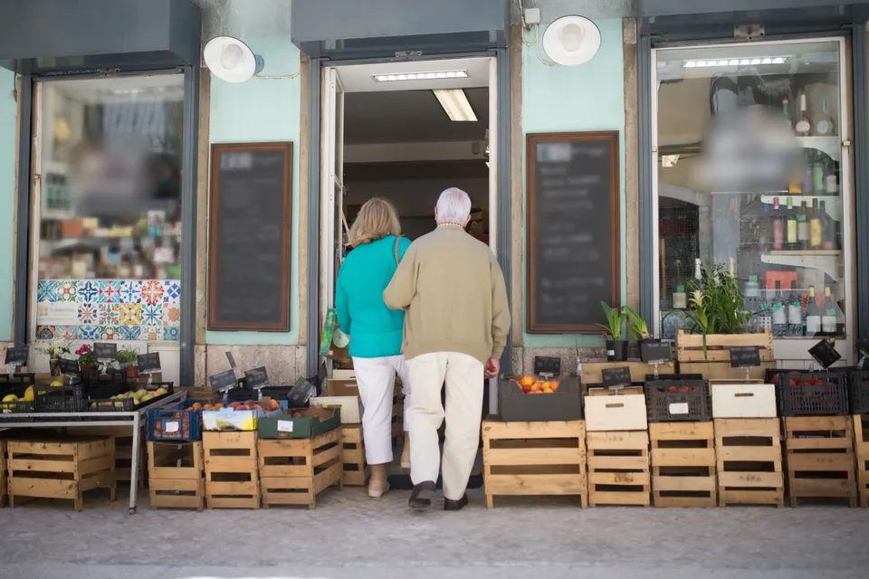 An older couple walking into a local grocery shop past crates of fresh produce.