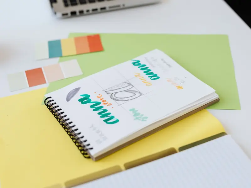 A spiral notebook of hand-lettered logo sketches next to paper colour swatches