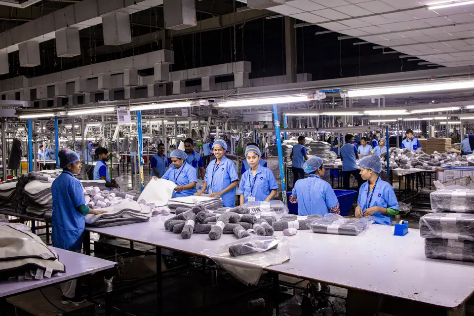 Rows of workers in blue uniforms and hairnets packing goods at long tables in a factory.