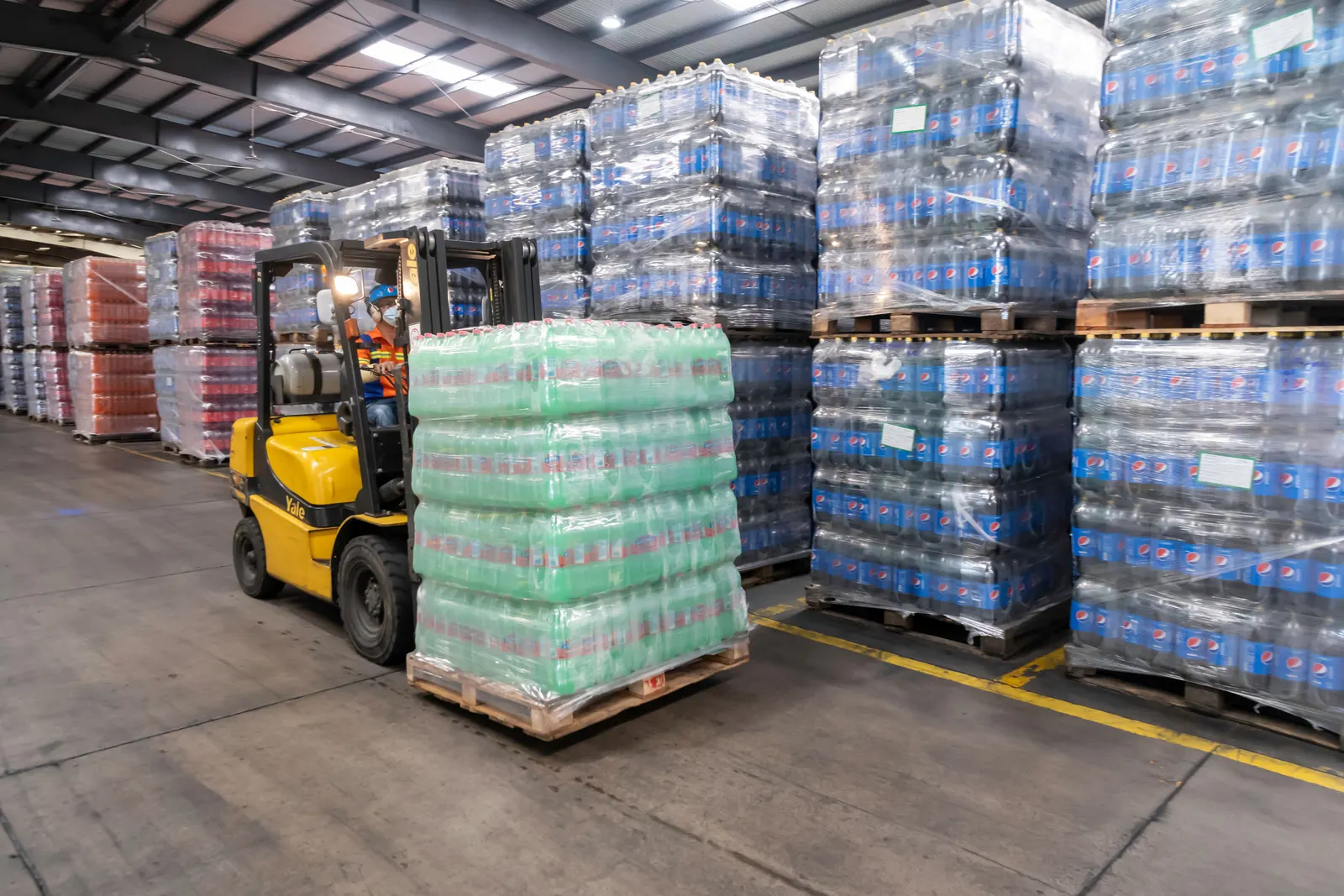 A forklift operator moving stacked pallets through a busy warehouse.